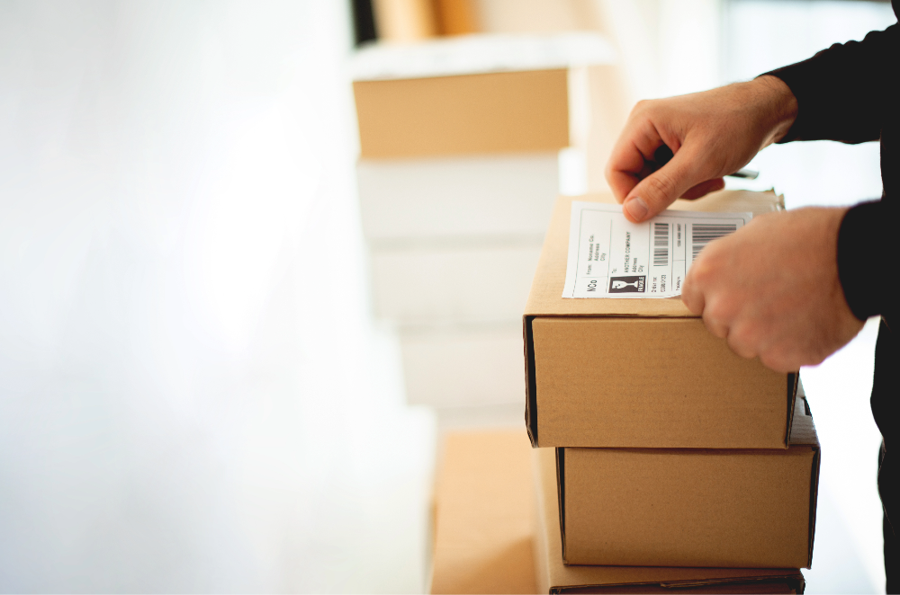 Person sealing a cardboard package with a shipping label, with more boxes stacked in the background.